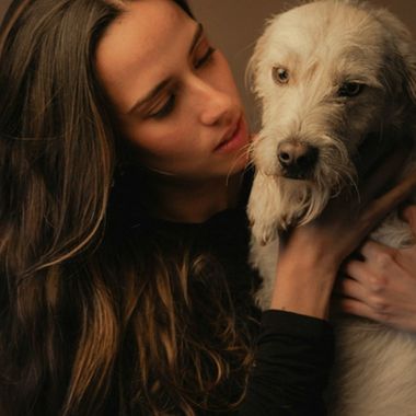 A person in a dark top cuddles a small white-and-tan dog with long fur, holding it close as they look down with a gentle expression.