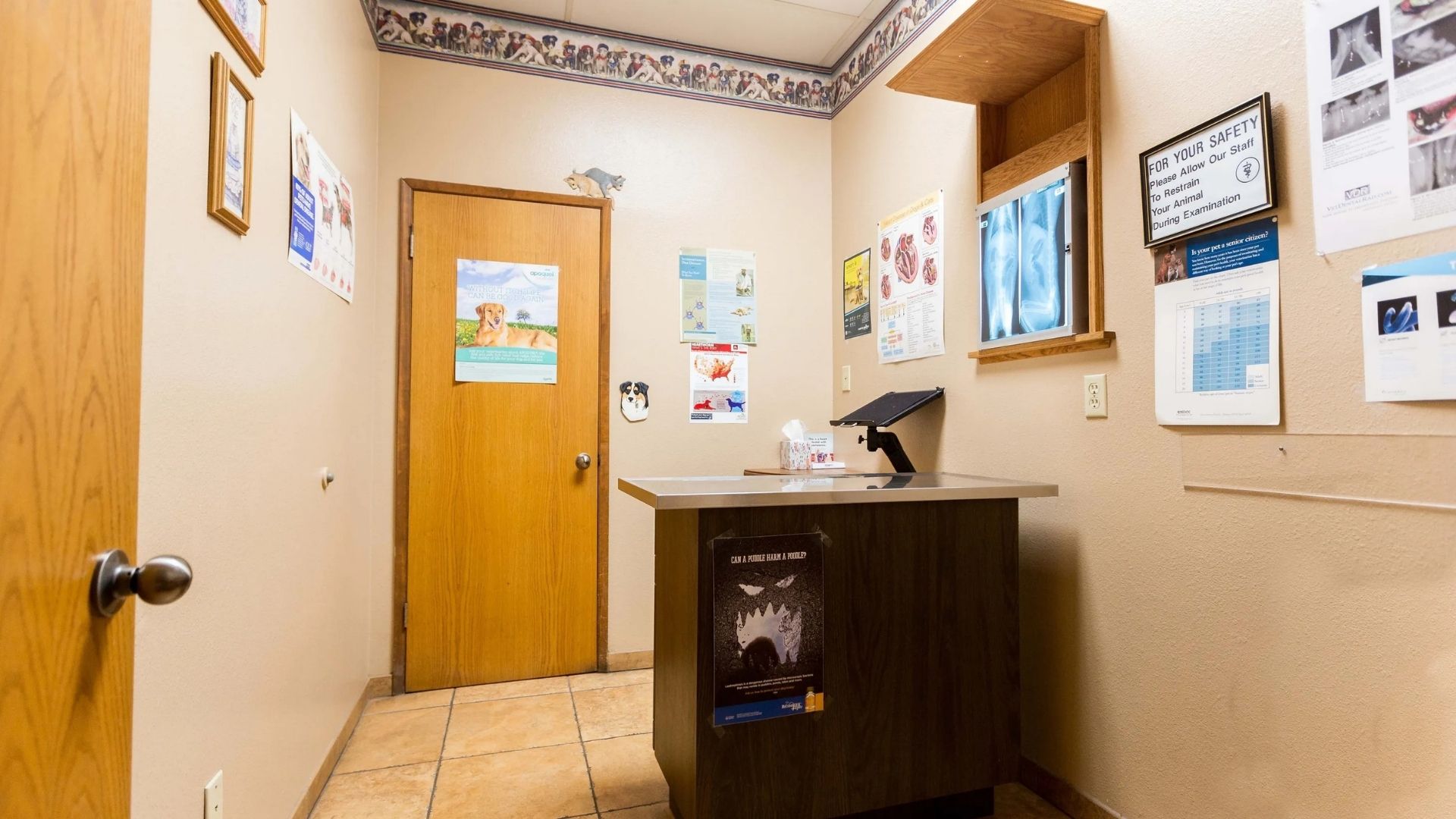 Reception Small office hallway with wooden door, bulletin boards and posters on cream walls, a calendar, and a dark wooden reception desk with a computer monitor, under fluorescent ceiling lights.
