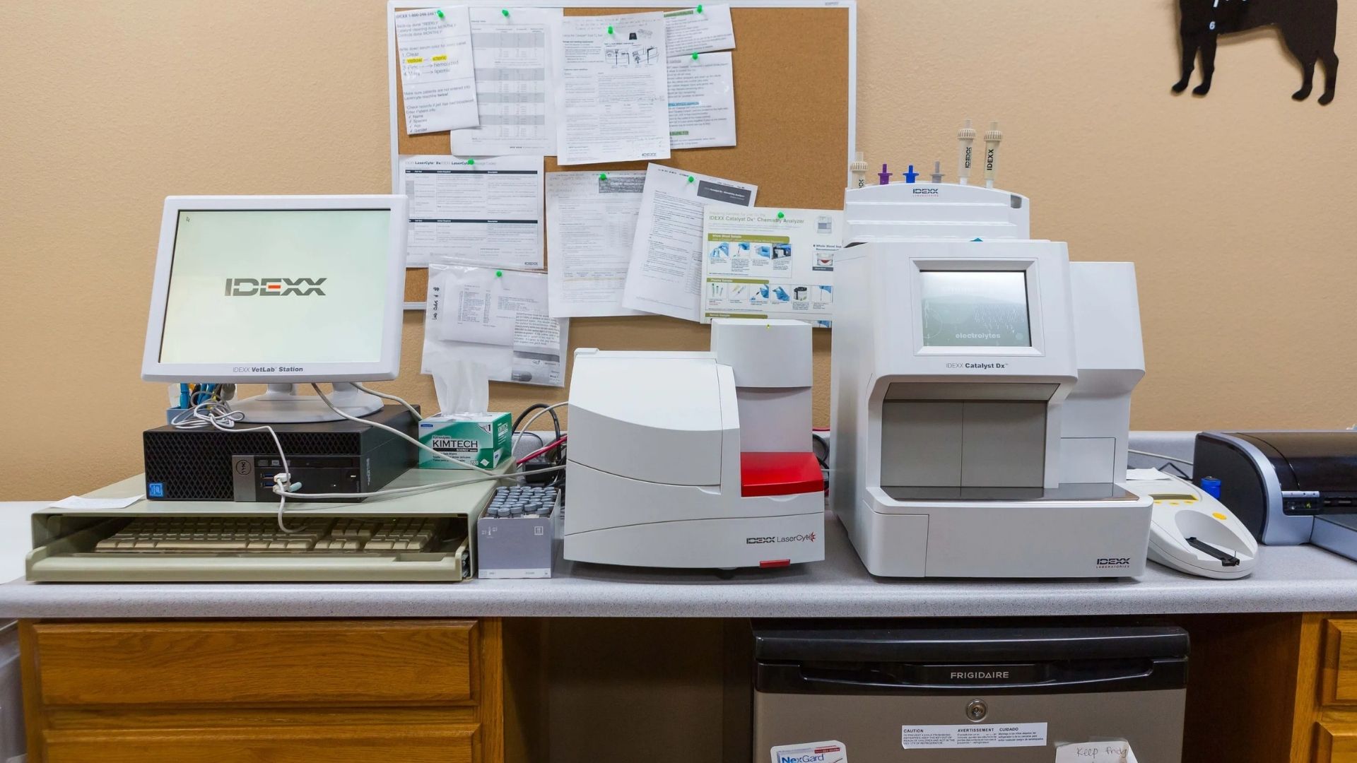 Table full of accessories Lab workbench with computer monitors, printed documents pinned on a board, and several measuring instruments and devices, including a white analyzer and a keyboard, on a wooden cabinet against a beige wall.